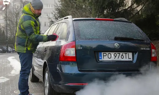 Mężczyzna w zimowej kurtce czyści zamarzniętą szybę auta. Dym z rury wydechowej sugeruje, że to diesel i właśnie próbuje się dowiedzieć, jak odpalić diesla w mroźny dzień.