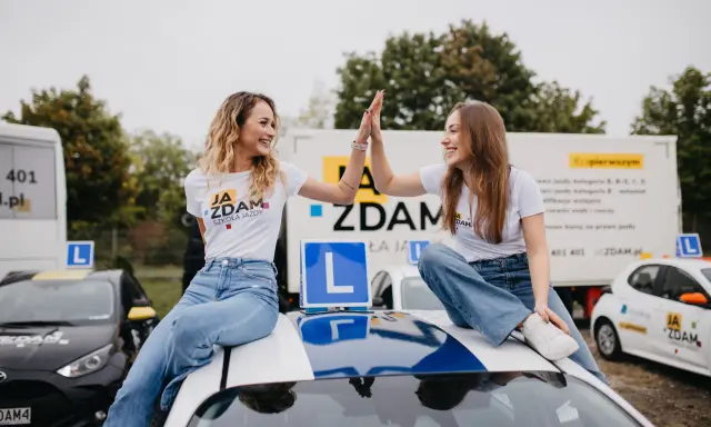 Dwie uśmiechnięte dziewczyny w koszulkach "JA ZDAM SZKOŁA JAZDY" dają sobie piątkę, siedząc na dachu samochodu szkoleniowego.
