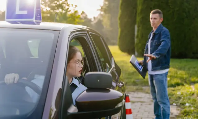 Młoda kobieta w okularach za kierownicą, z literą "L" na dachu, uczy się jeździć. Instruktor z tablicą daje wskazówki. To jej kontynuacja prawa jazdy po latach.