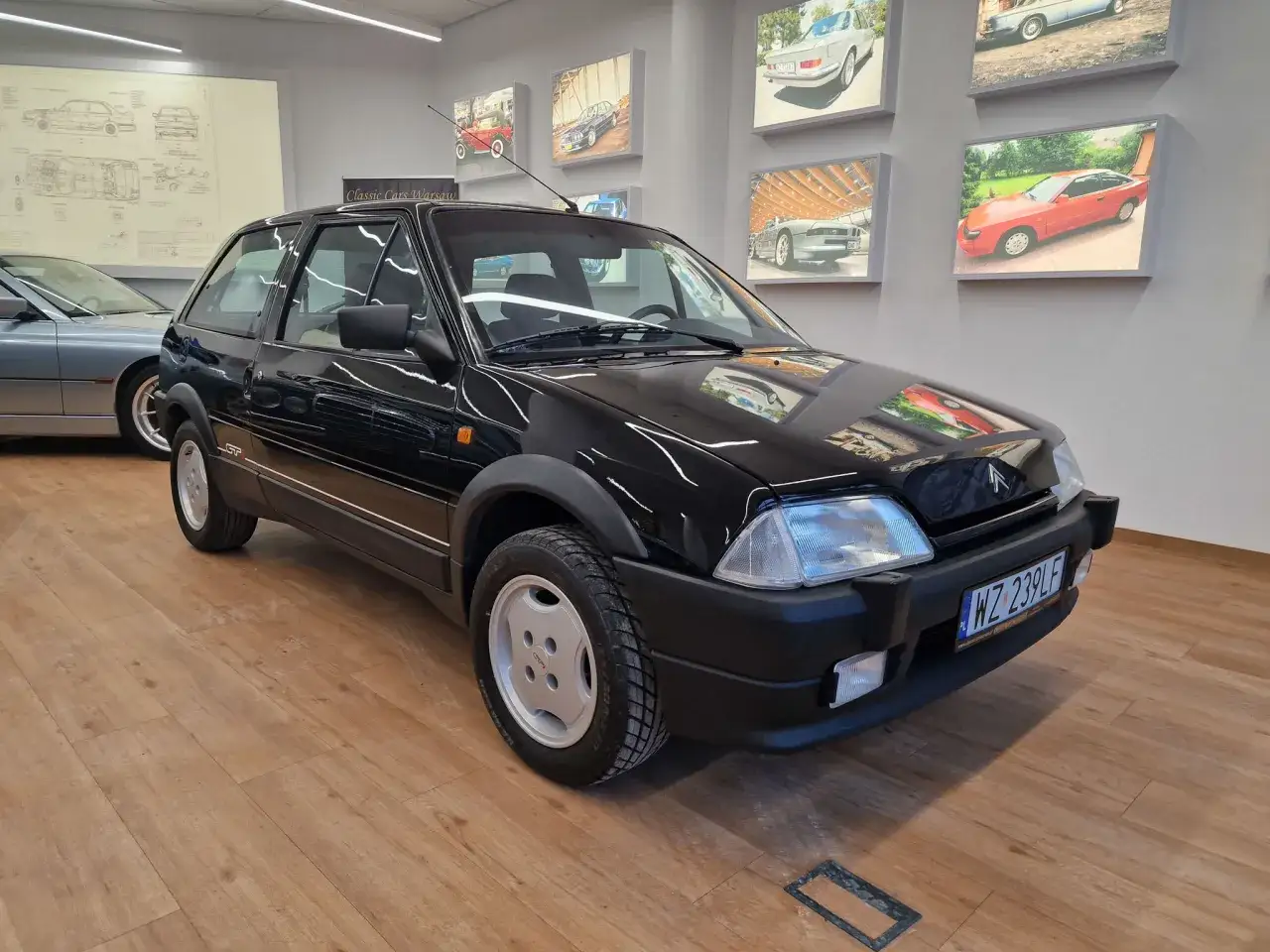 Czarny Citroen AX, jeden z klasycznych modeli, prezentuje się na drewnianej podłodze. W tle wiszą zdjęcia innych zabytkowych samochodów.