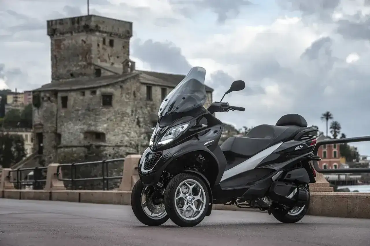 Czarny Piaggio MP3 500 z imponującą prędkością maksymalną, zaparkowany przed zabytkową wieżą nad morzem.