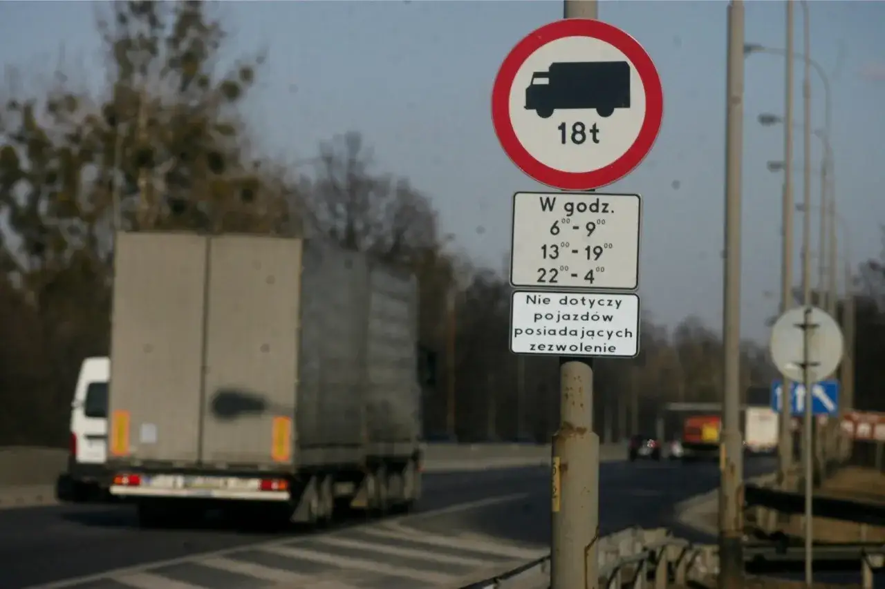 Znak zakazu wjazdu dla ciężarówek powyżej 18t w określonych godzinach. Czy ciężarówki mogą jeździć w niedzielę? Zależy od zezwolenia.