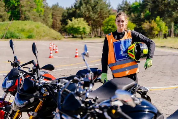 Młoda kobieta w kamizelce z literą "L" i kaskiem w ręku, gotowa do jazdy na motocyklu na placu manewrowym. To jej pierwsze kroki na drodze, a wkrótce będzie mogła jeździć na A1.