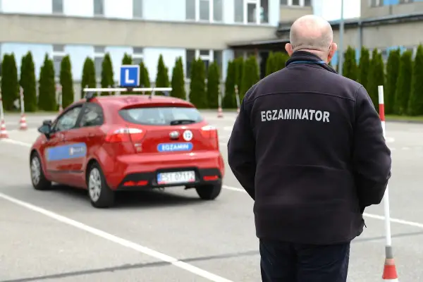 Egzaminator w czarnej kurtce z napisem "EGZAMINATOR" obserwuje czerwony samochód z literą "L" na dachu. Jak odwołać egzamin na prawo jazdy infocar?